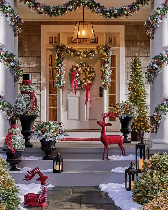 Grand white porch with snow, red reindeer figures, wreaths, and glowing lanterns