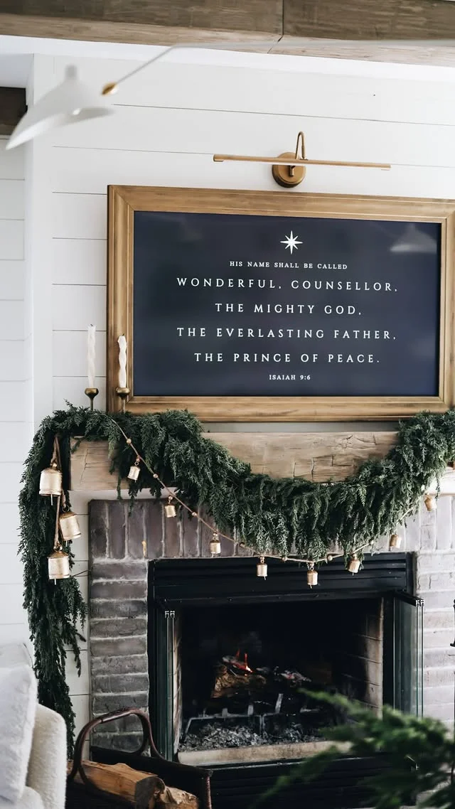 White mantel with layered green garland, brass bells, and framed holiday art