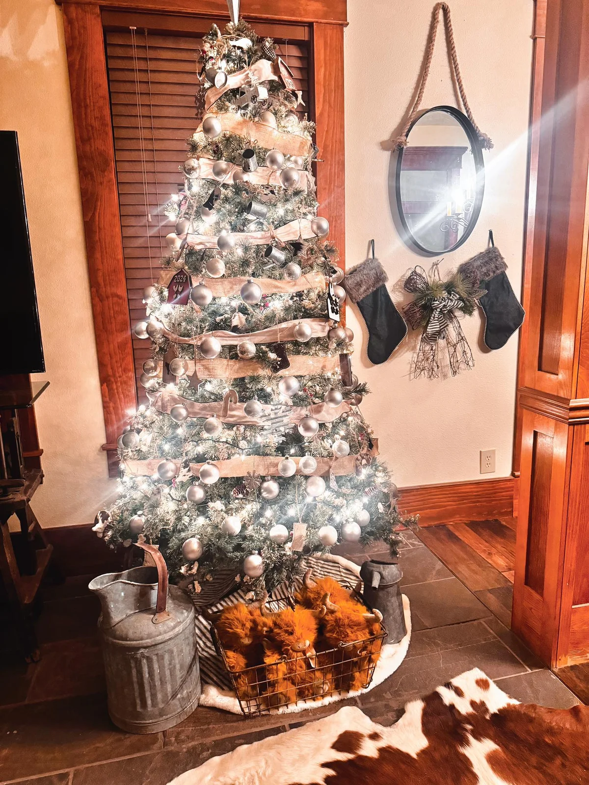 Christmas tree with silver ornaments and burlap ribbon glowing in front of a window