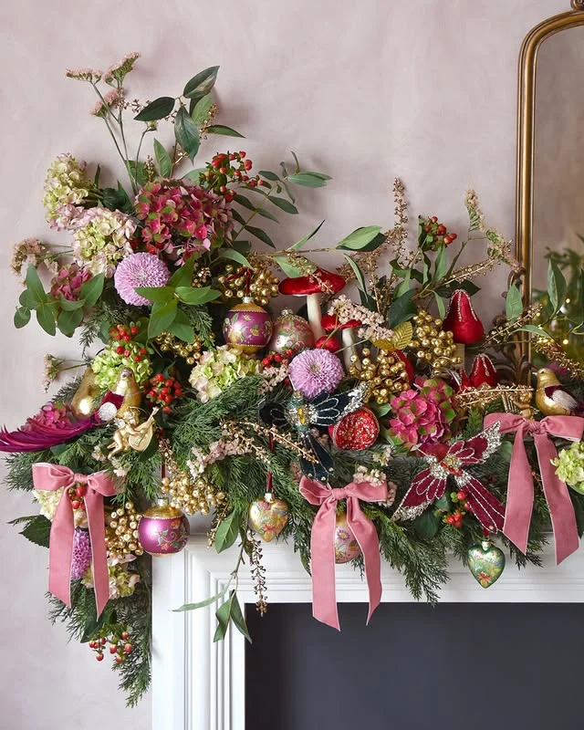 White fireplace mantel decorated with a lush Christmas garland of greenery, pink flowers and pink velvet bows